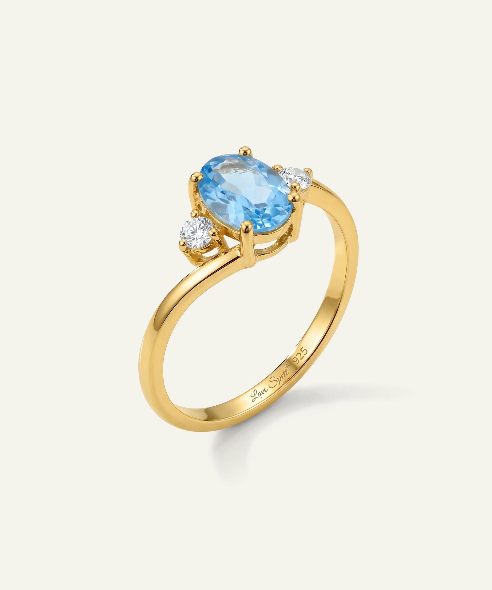 A yellow gold ring featuring an oval-cut light blue topaz center stone and two small round-cut clear accent stones, shown on a clean off-white background. The inner band is engraved with 'Jewelry by Jane'.