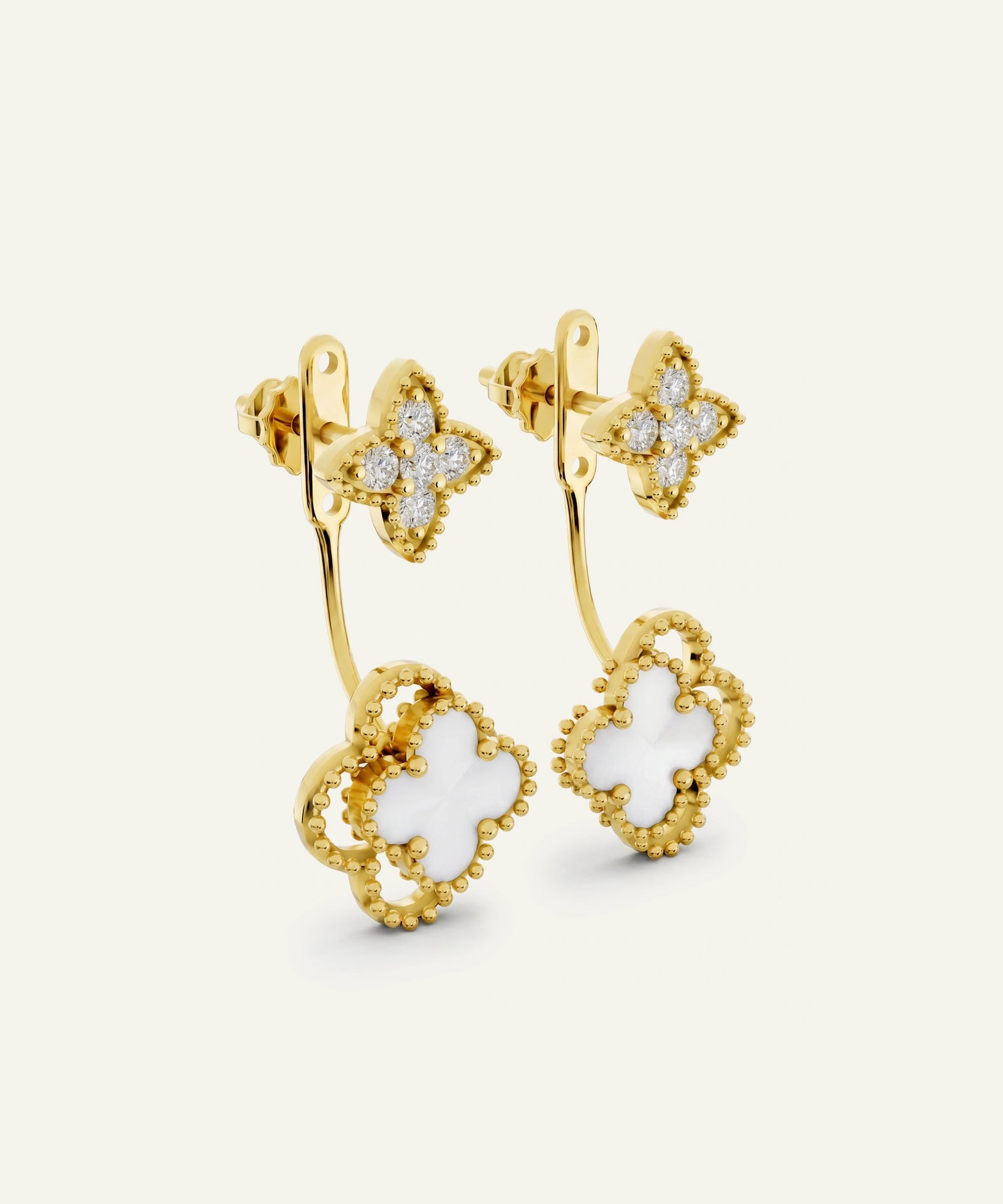 A pair of gold-toned huggie hoop earrings featuring a double-layered Alhambra-style quatrefoil drop charm with a white inlay and beaded gold borders, accented with small clear stones on the upper hoop.