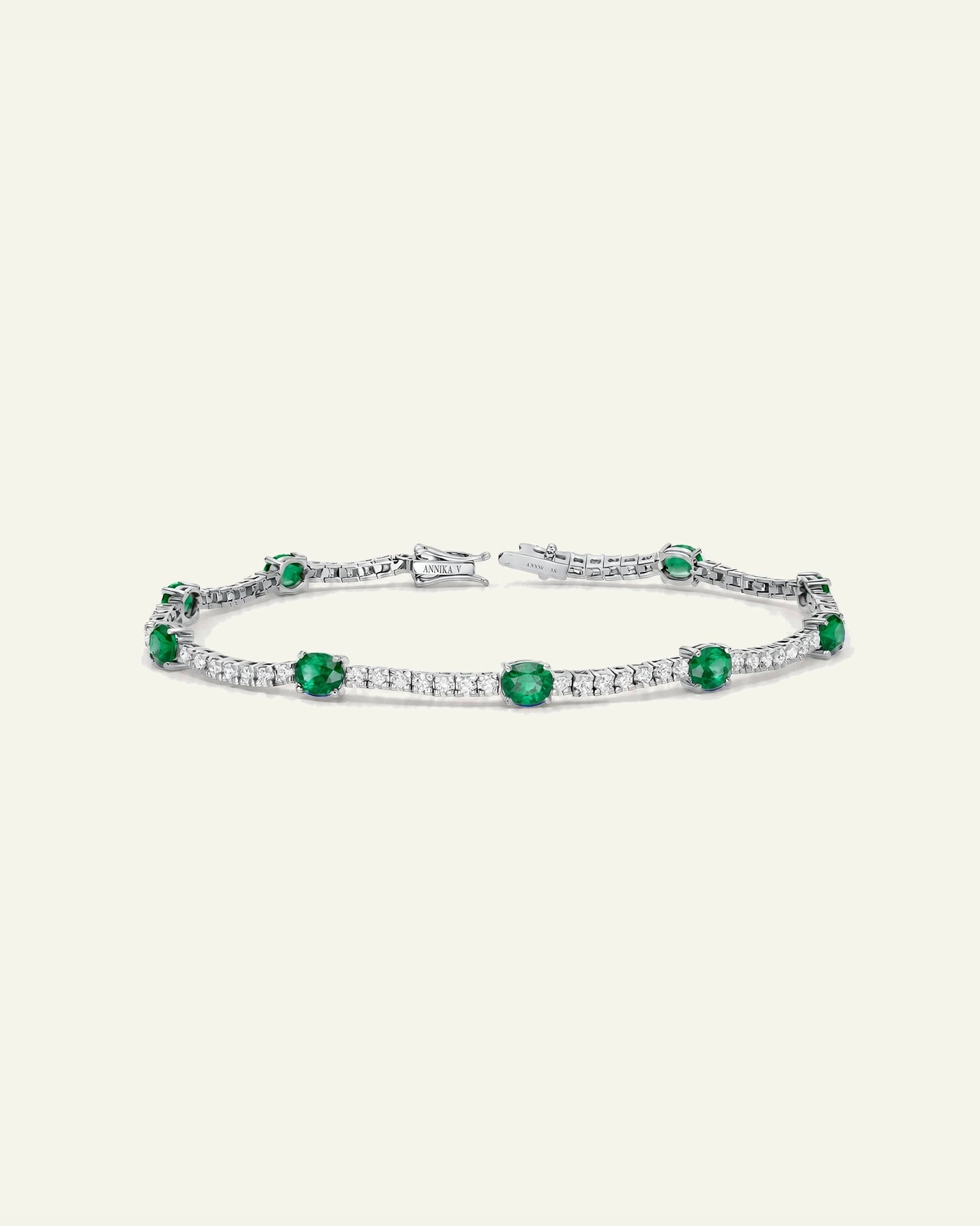 A silver tennis bracelet featuring round-cut green emeralds and pavé-set clear stones, displayed on a light cream background.
