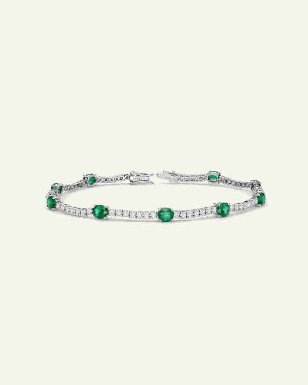 A silver tennis bracelet featuring round-cut green emeralds and pavé-set clear stones, displayed on a light cream background.