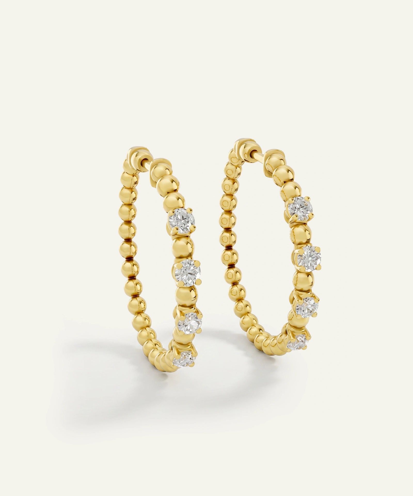 A pair of gold hoop earrings with a beaded texture and small clear round-cut stones, photographed on a clean off-white background.