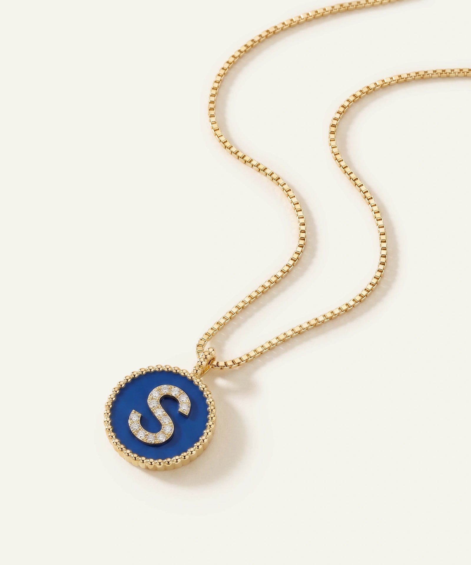 A gold chain necklace with a round pendant featuring a vibrant blue enamel background and a raised gold letter 'S' outlined with small clear stones, set against a clean off-white background.