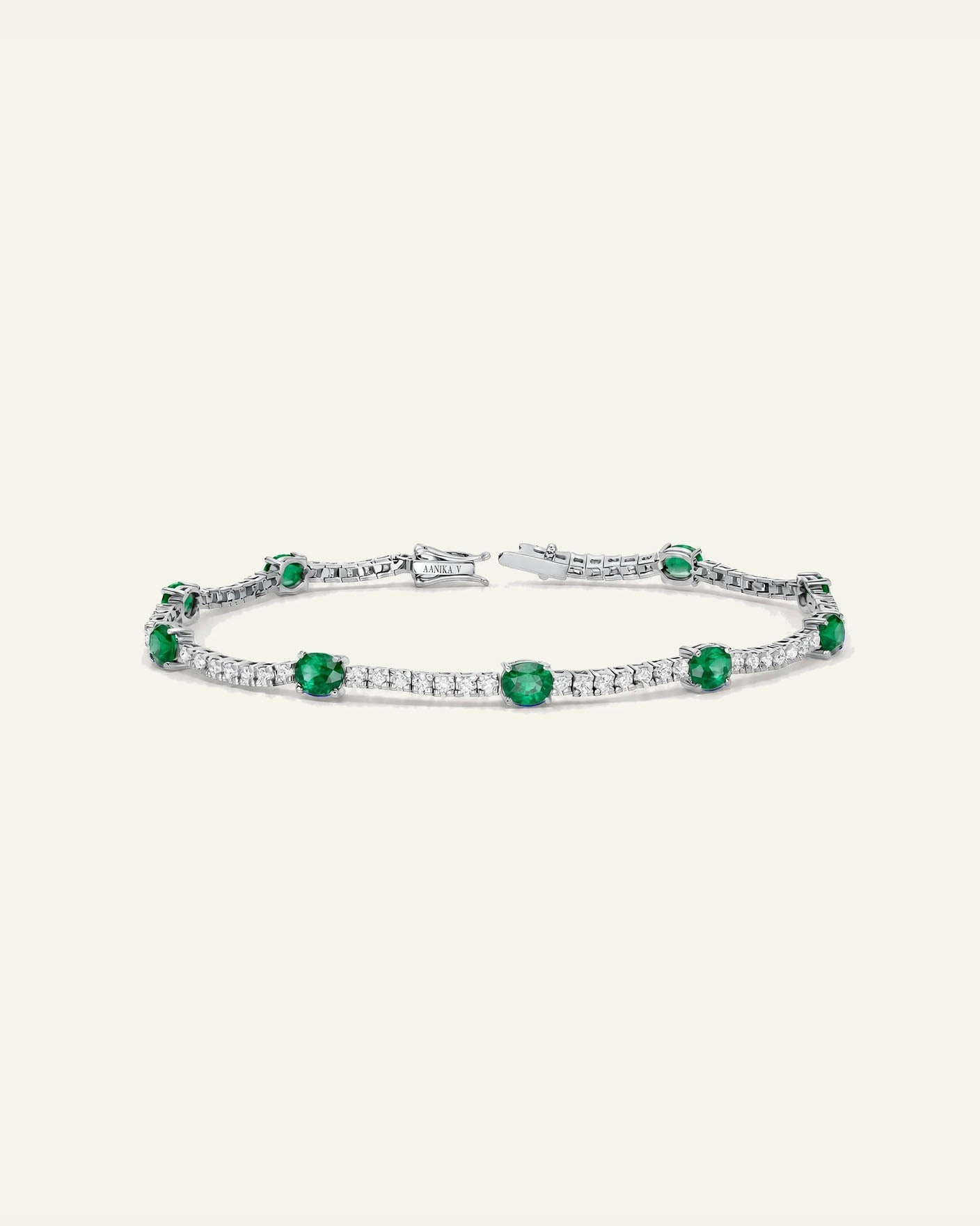 A silver tennis bracelet featuring round-cut green emeralds and pavé-set clear stones, displayed on a light cream background.