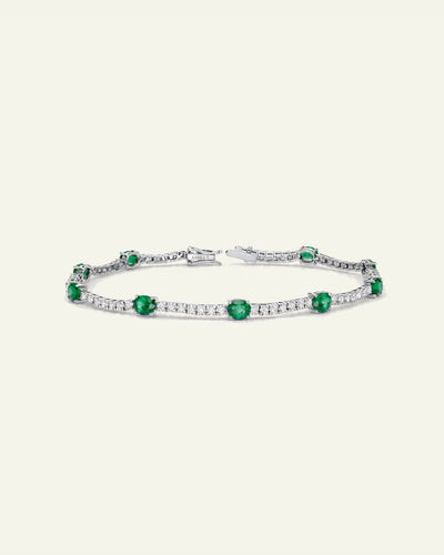 A silver tennis bracelet featuring round-cut green emeralds and pavé-set clear stones, displayed on a light cream background. - alternate view