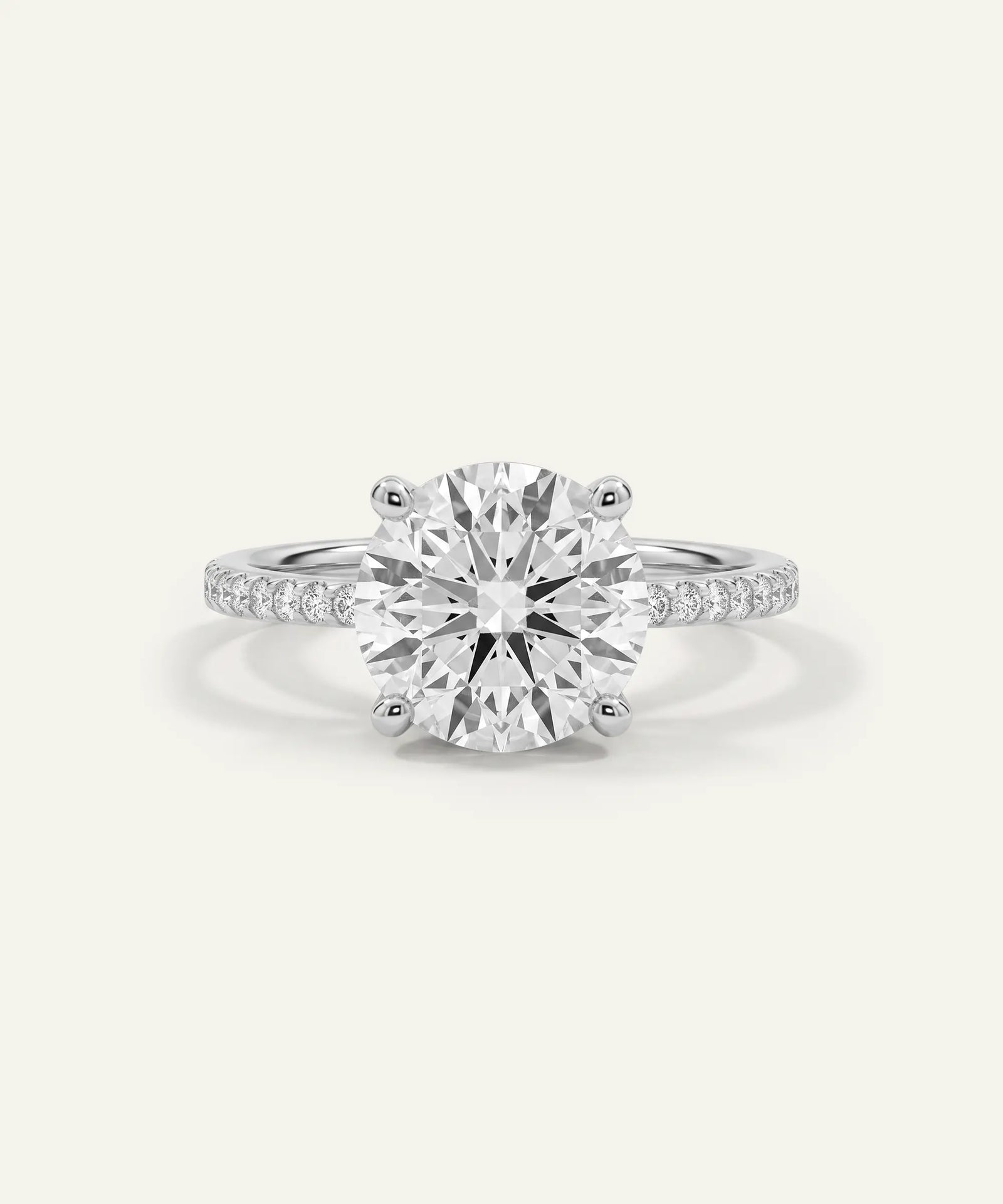 A classic solitaire engagement ring featuring a round brilliant-cut diamond center stone in a six-prong setting, with a pavé band set with small round diamonds, shown on a light cream background.