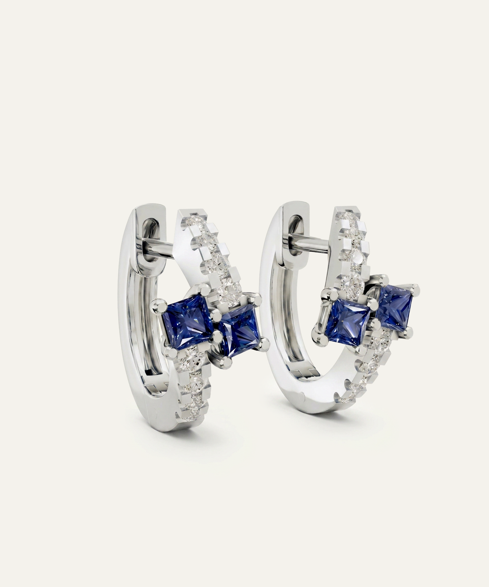 A pair of silver huggie hoop earrings featuring square-cut blue sapphires and pavé-set clear crystals, shown on a light off-white background.