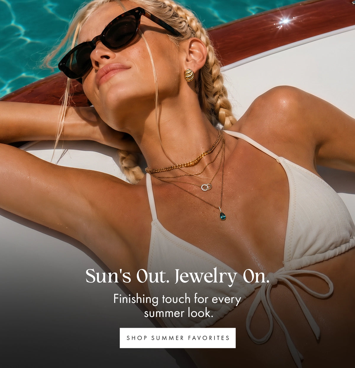 A woman reclining by a pool wearing black sunglasses, layered gold necklaces, and a white bikini top, with promotional text reading 'Sun's Out. Jewelry On.