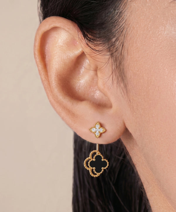 A close-up of a model's ear wearing a gold drop earring with a small pavé diamond four-petal stud and a dangling gold quatrefoil with a black center.