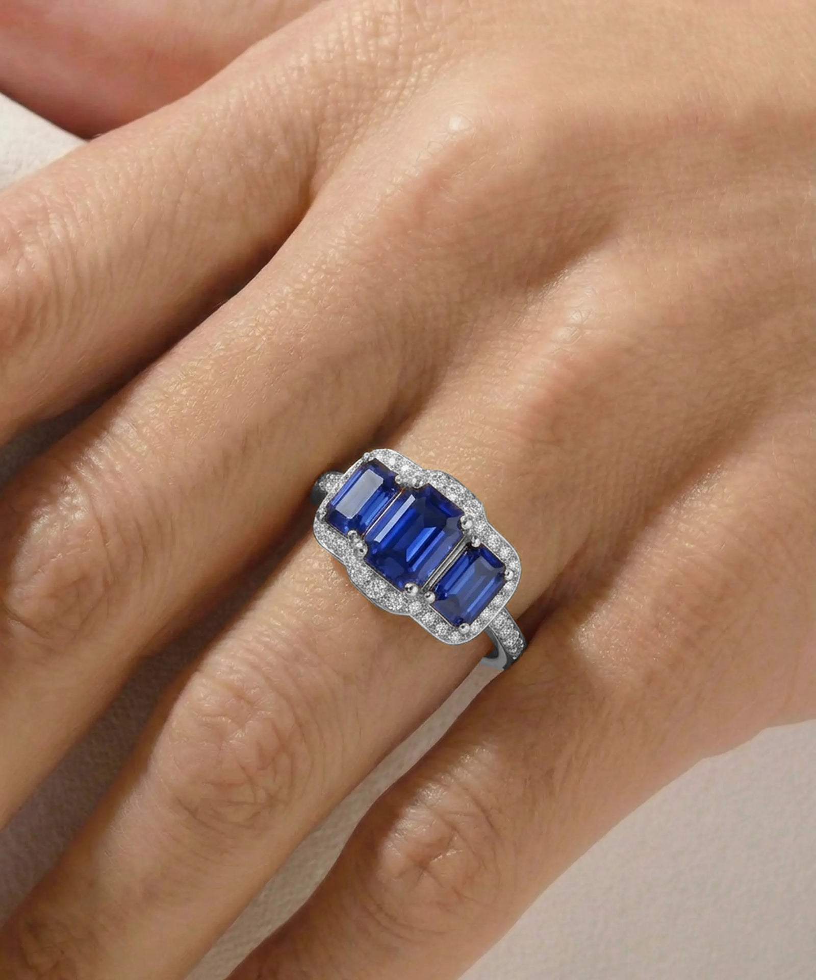A close-up of a hand wearing a silver ring with three emerald-cut blue sapphires and a halo of small clear stones.