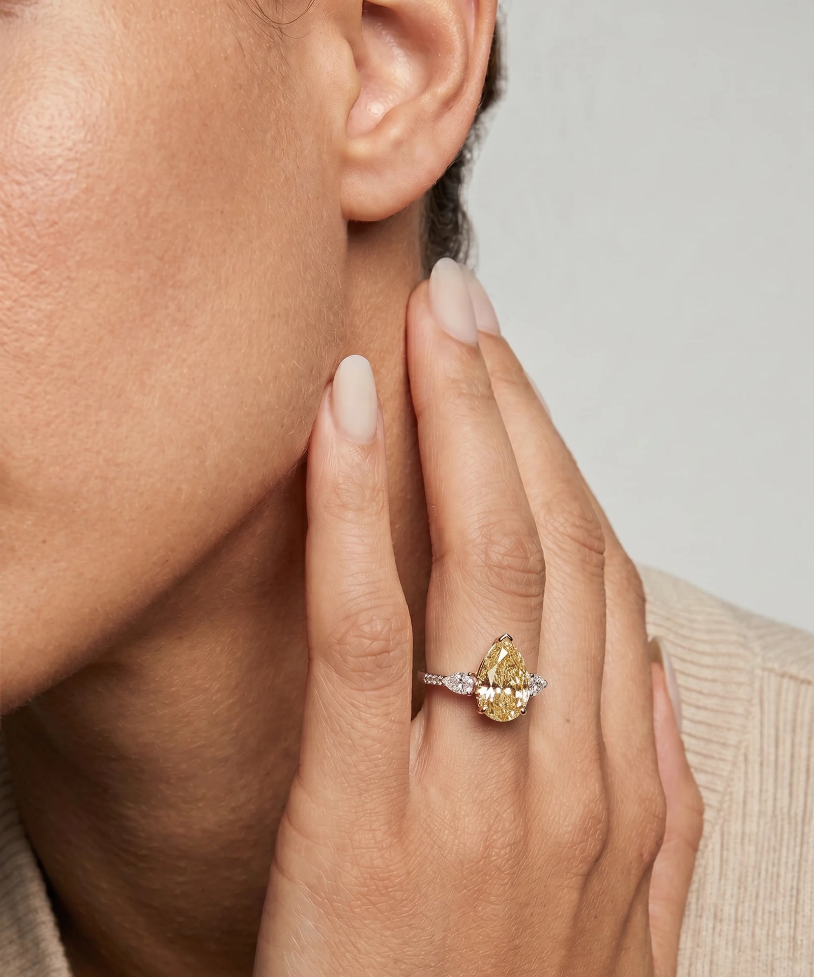 Close-up of a hand wearing a gold ring with a large gemstone, touching the ear against a neutral background.