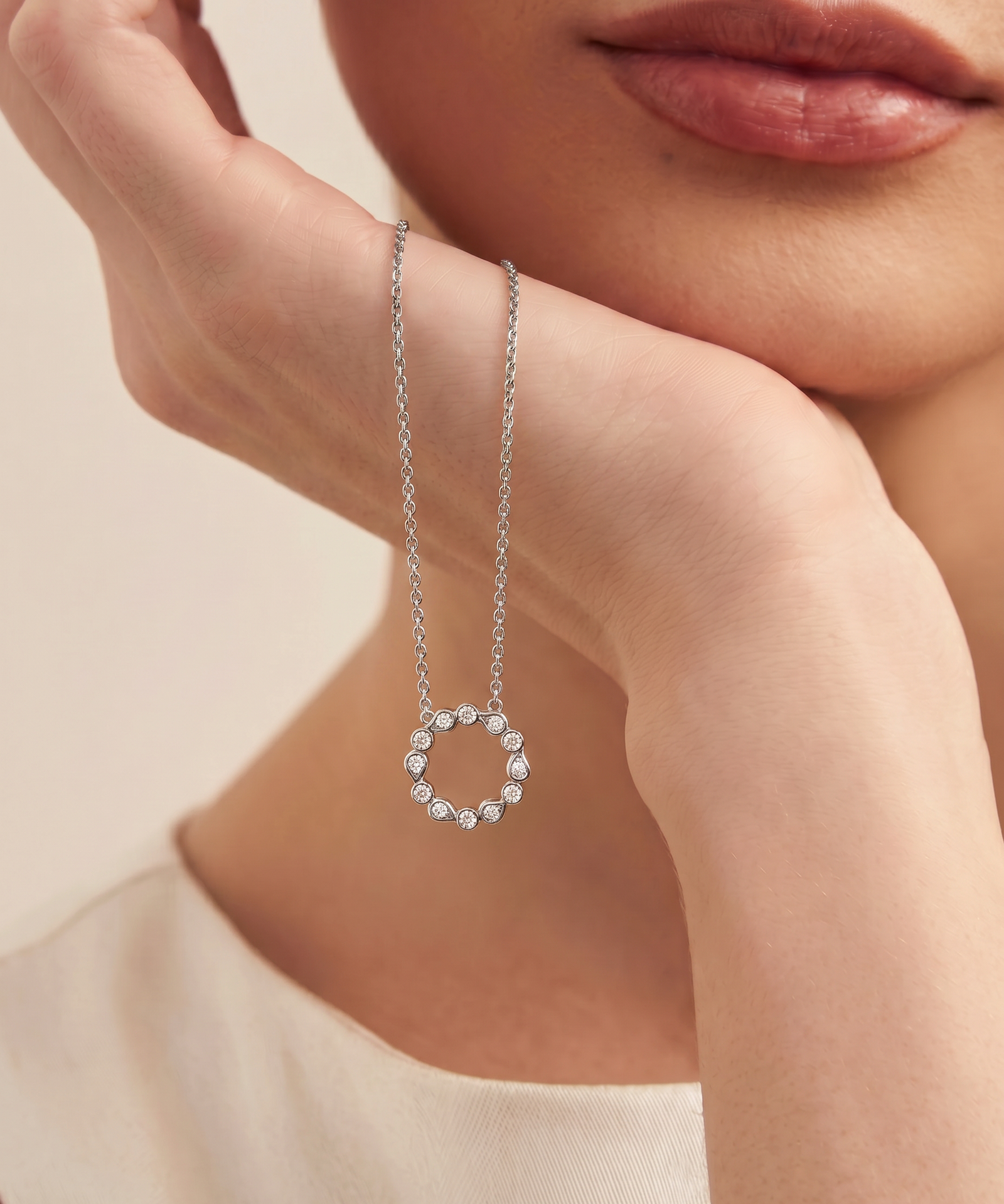 A close-up lifestyle shot of a model resting her chin on her hand, showcasing a delicate silver chain necklace with a small, open circular pendant adorned with tiny clear stones, set against a soft beige background.