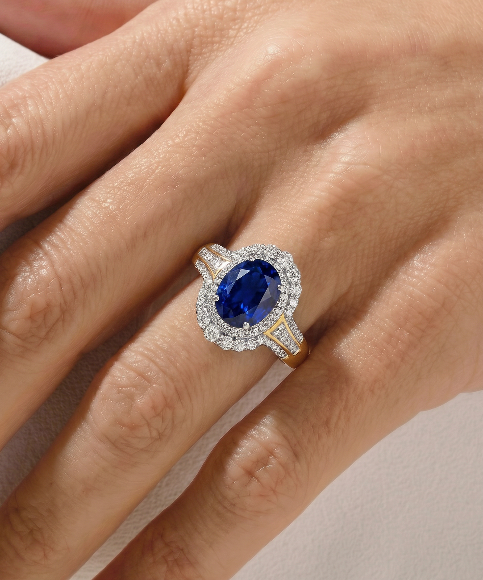 A close-up of a hand wearing a ring with an oval blue sapphire center stone surrounded by a halo of small clear stones, with a yellow gold band featuring small accent stones.