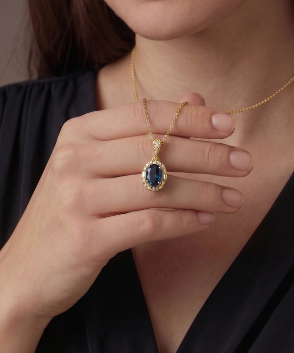 Gold necklace with a blue gemstone pendant held by a hand against a neutral background