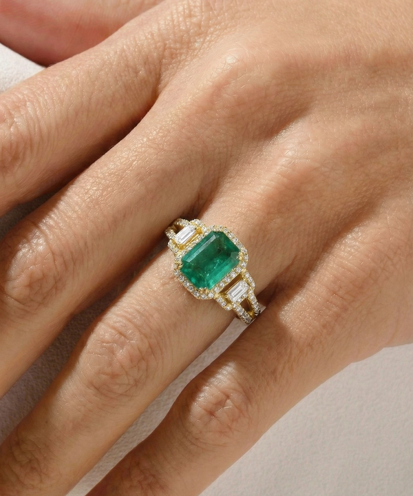Emerald ring with diamond accents on a hand against a neutral background
