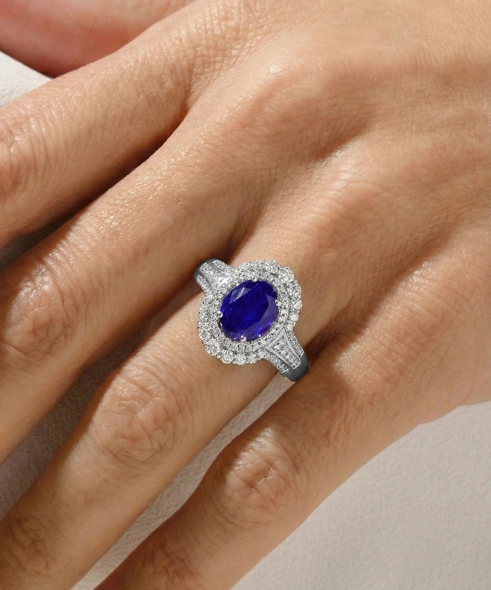 A close-up of a hand wearing a silver-toned halo ring with a large oval deep blue gemstone and small clear accent stones.