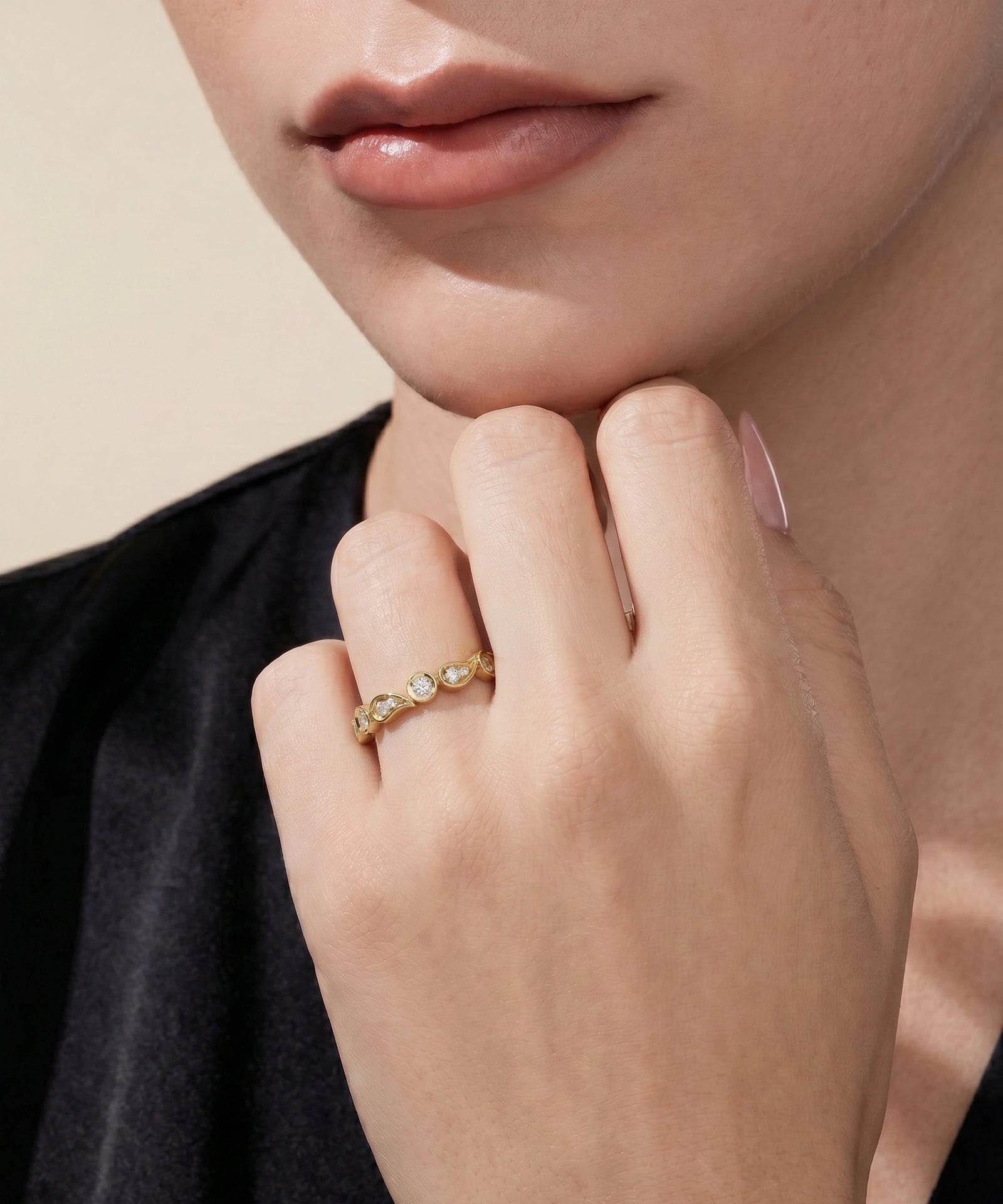 A close-up of a model's hand wearing a delicate gold ring with small clear stones, set against a soft beige background and black top.