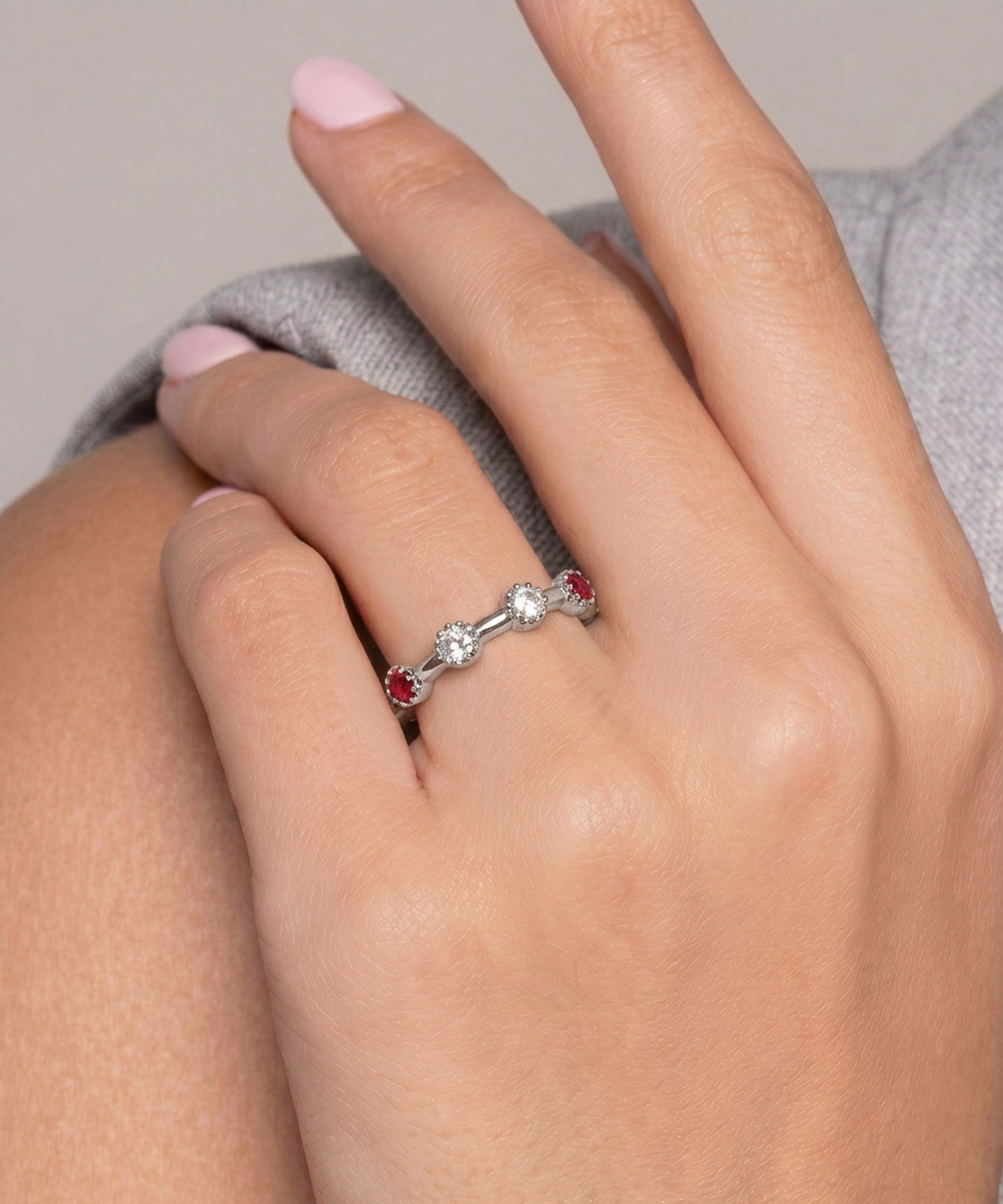 A close-up of a hand wearing a silver ring with three small round stones, including two red stones and one clear center stone, resting on a gray fabric surface.