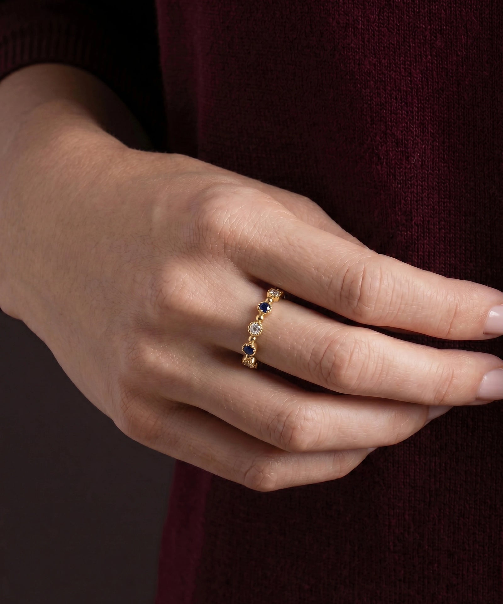 A close-up of a person's hand wearing a delicate gold band ring with small dark blue and clear gemstones, set against a dark burgundy knit sweater.