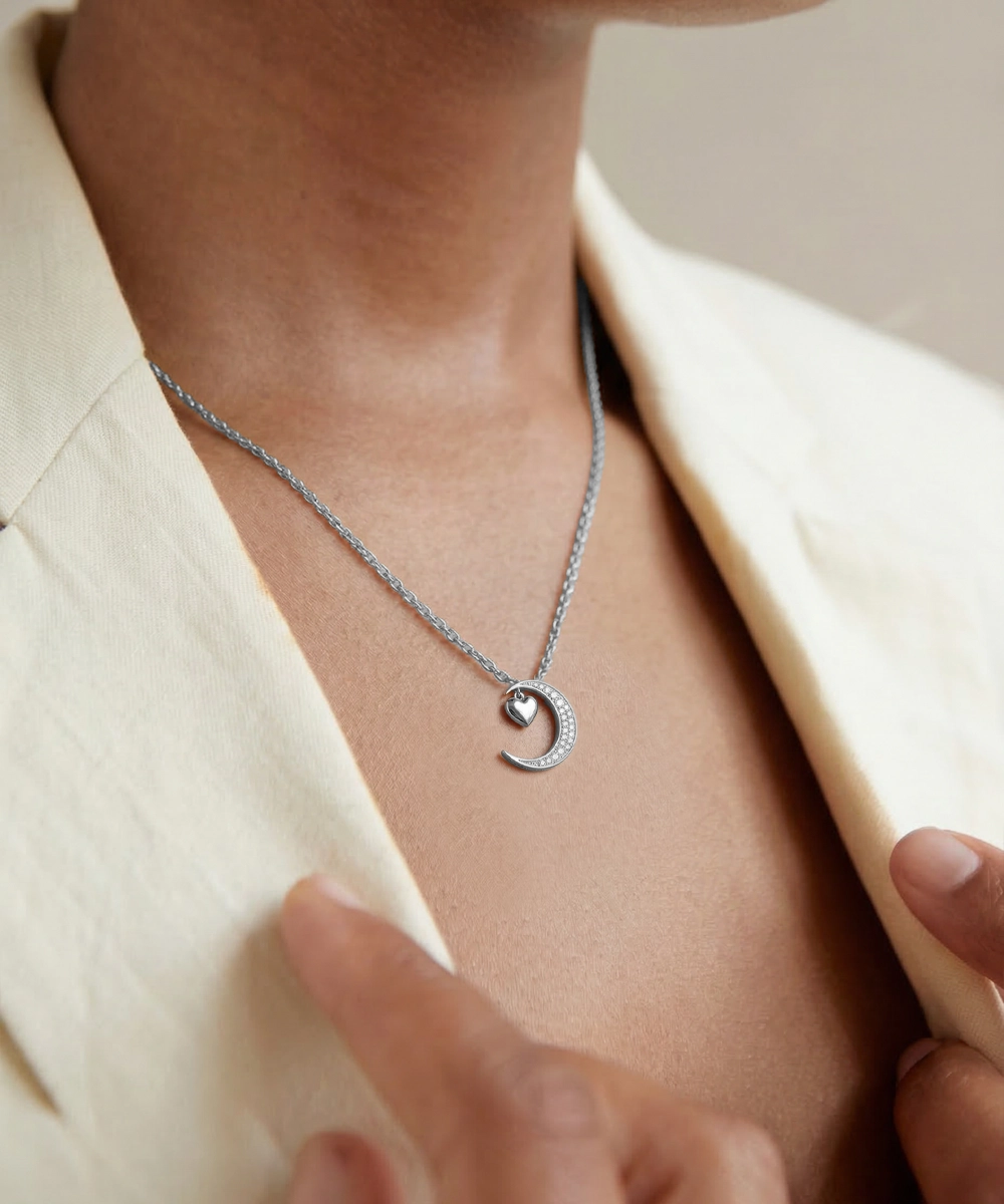 Silver necklace with a heart and crescent moon pendant worn by a person in a beige outfit.