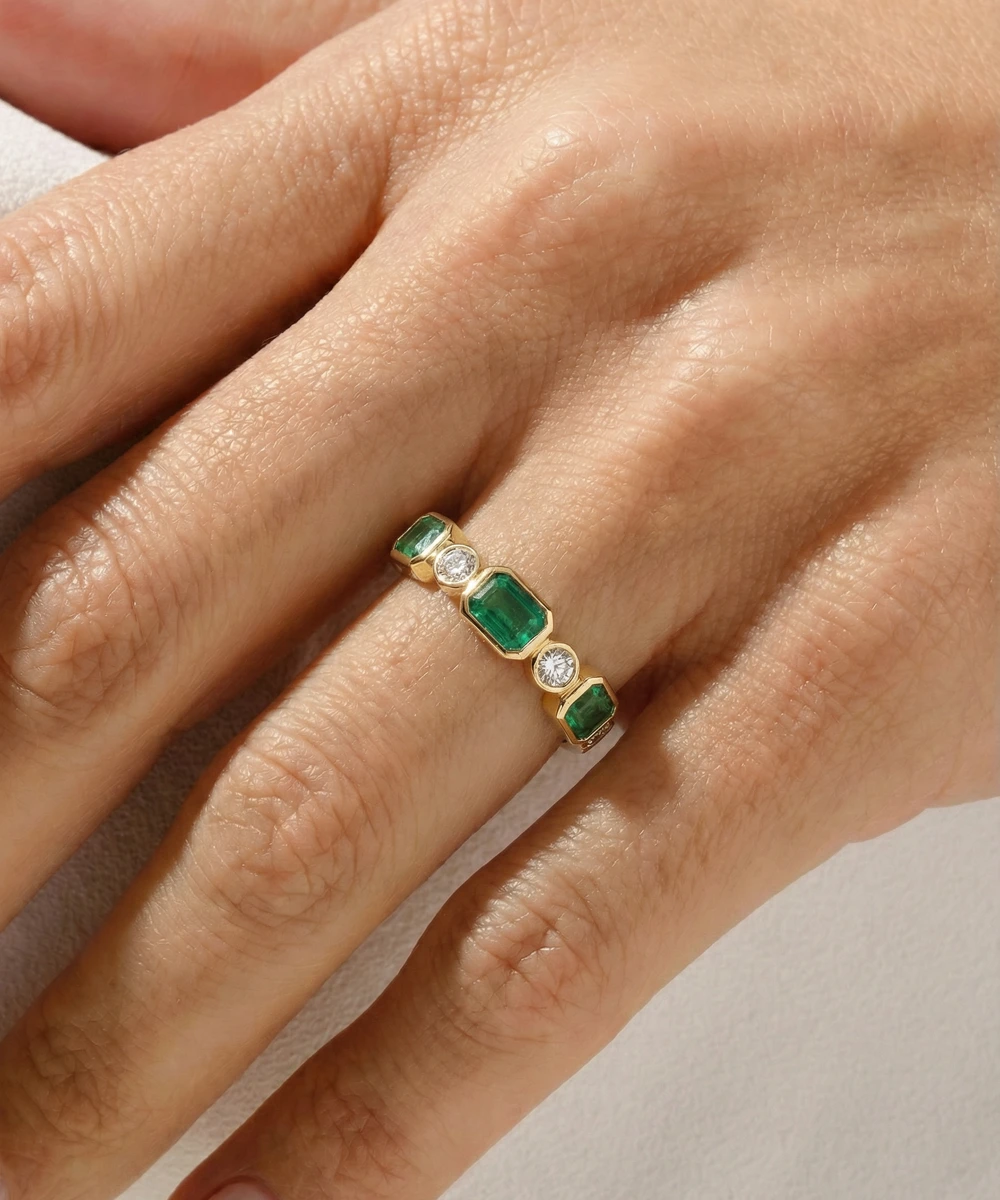 Gold ring with emerald and diamond stones on a hand against a neutral background