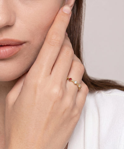 A close-up lifestyle shot of a model wearing a delicate gold ring on her finger. The ring features a small red gemstone on one side and a white pearl on the other, set against a soft, light gray background. - alternate view