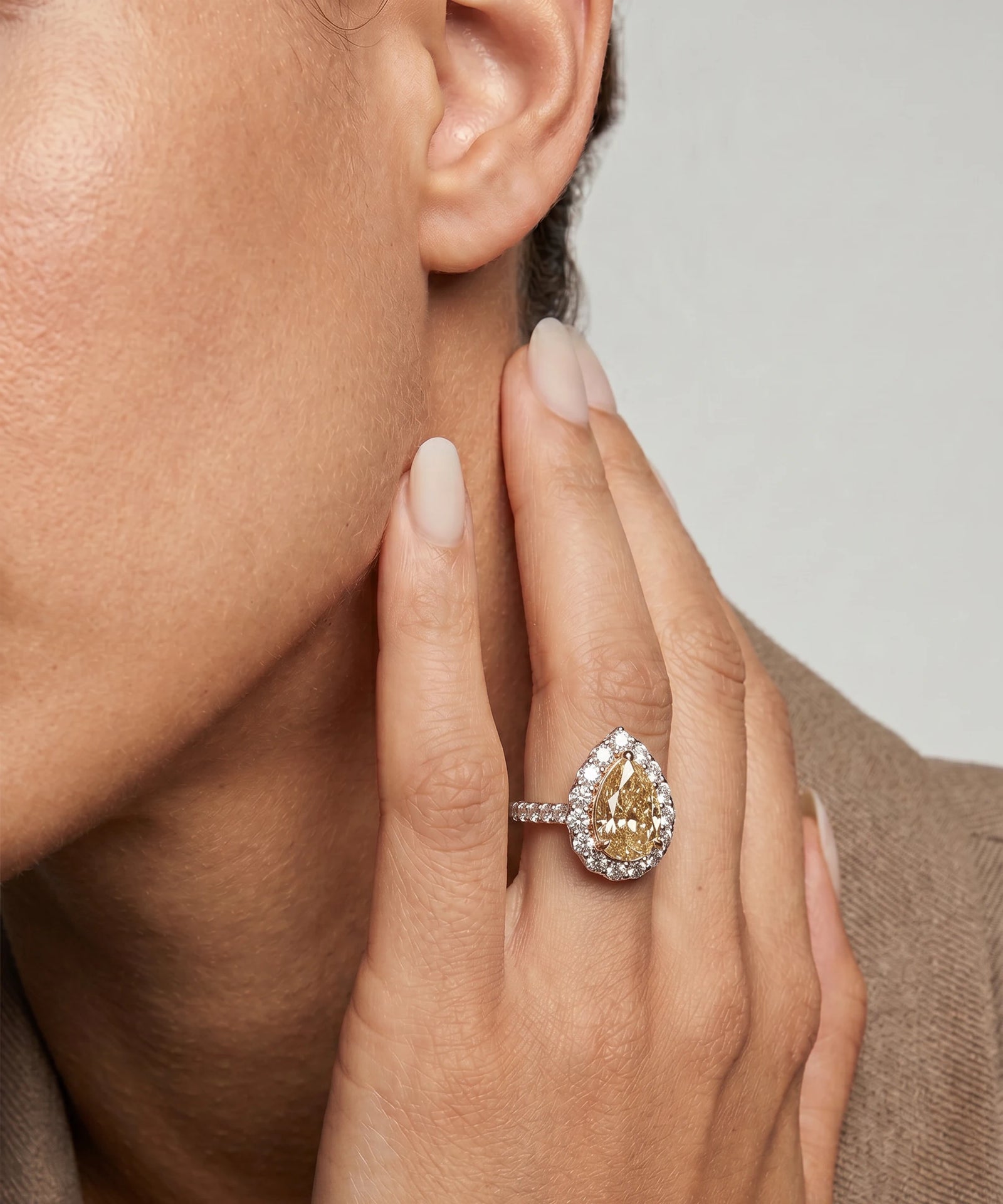 Close-up of a hand wearing a diamond ring with a light brown gemstone, held near an ear.