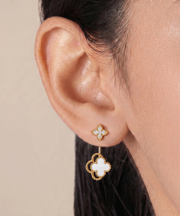 A close-up of a model's ear wearing a gold and white clover drop earring against a soft beige background.