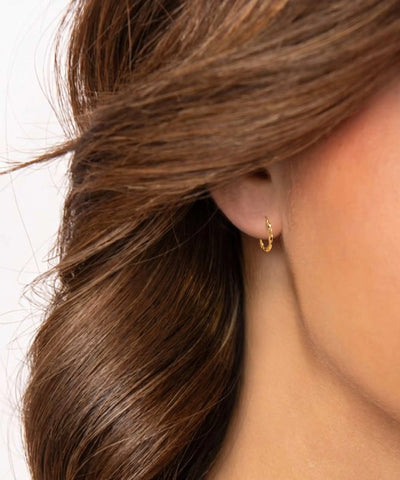 A close-up of a model's ear wearing a small gold huggie hoop earring, with brown hair framing the side of the face against a plain light background. - alternate view