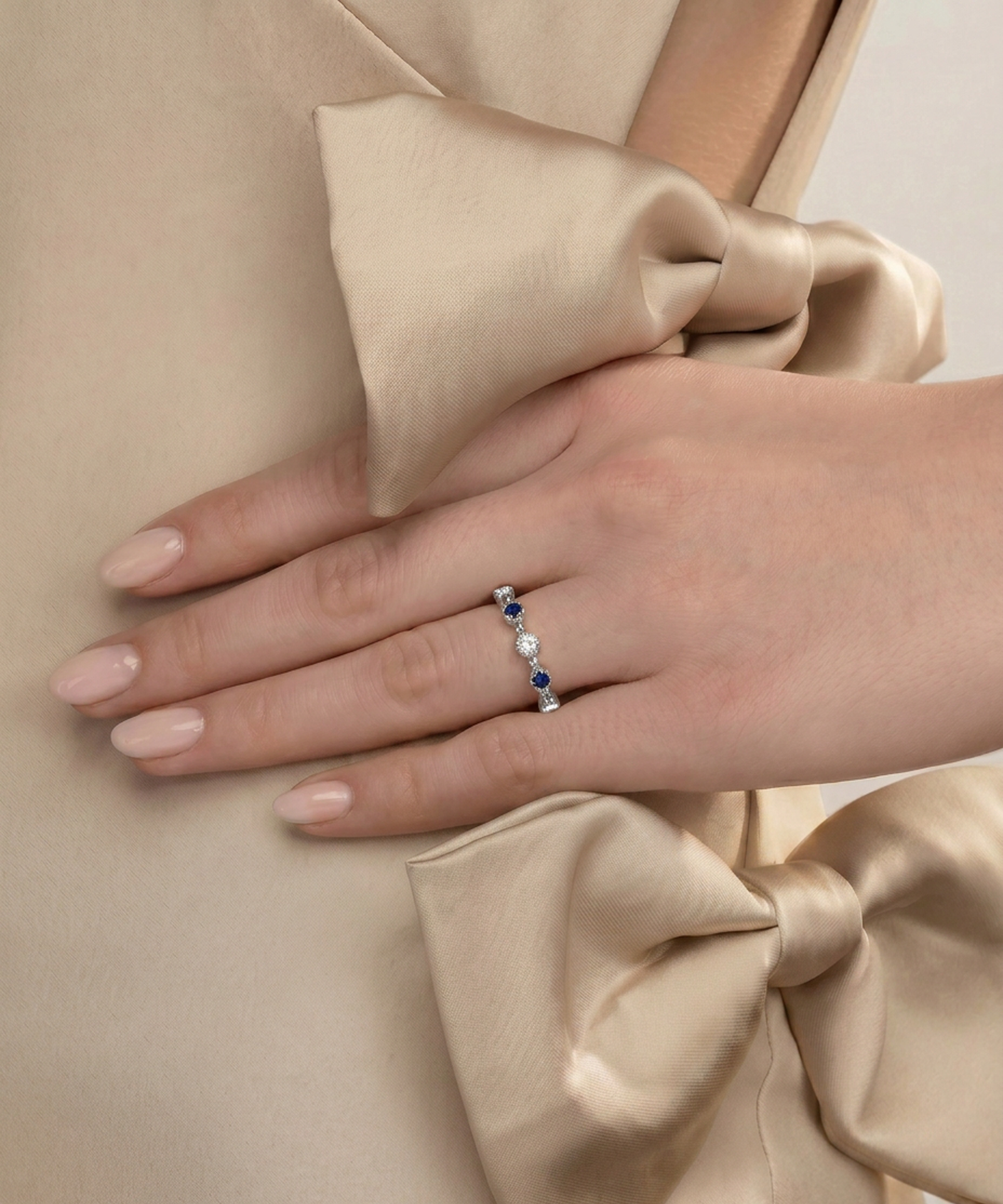 A close-up of a hand wearing a delicate silver ring with small blue and white stones, resting on a beige satin dress with a large bow.