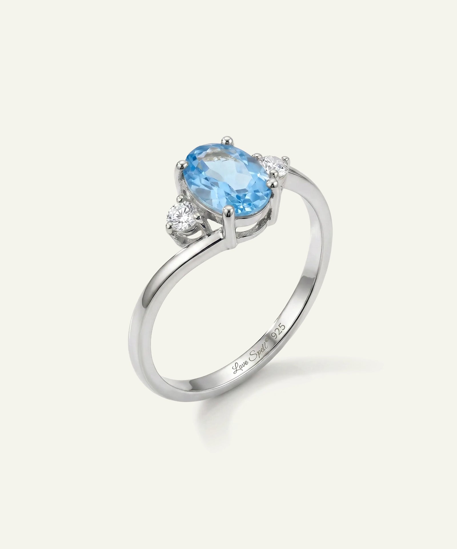 A silver ring featuring an oval-cut light blue gemstone in a prong setting, accented by two small round clear stones on either side, shown on a light cream background.