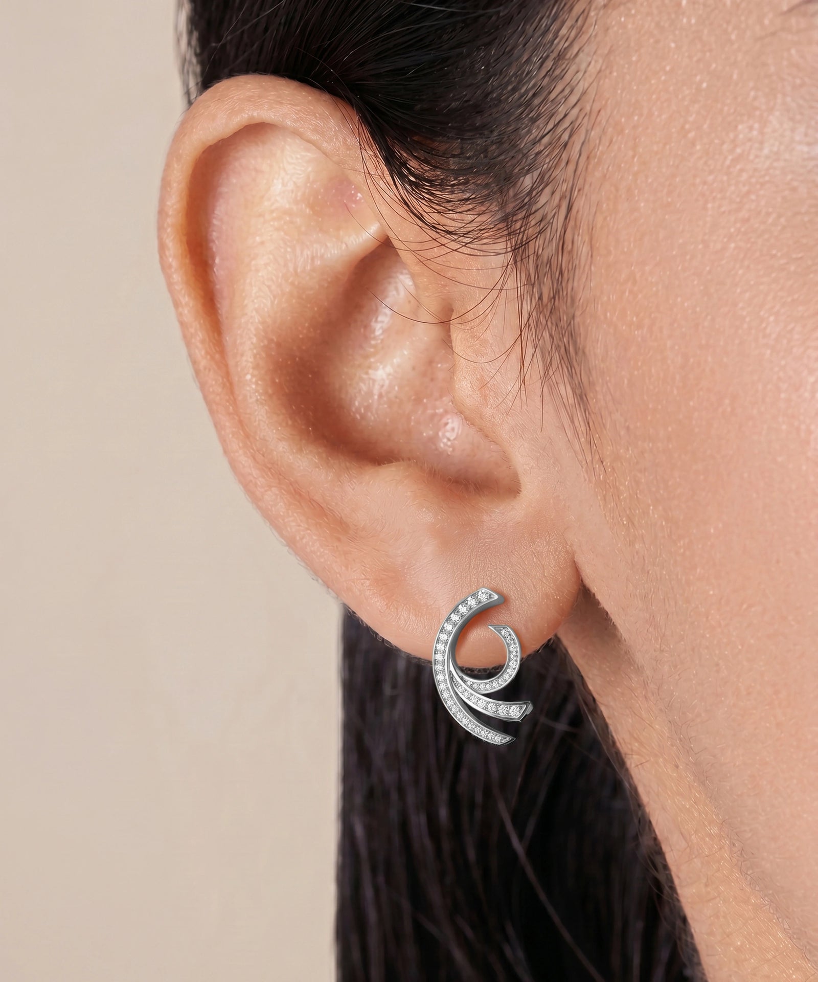 A close-up of a model's ear wearing a silver crescent-shaped stud earring set with small clear stones, against a soft beige background.