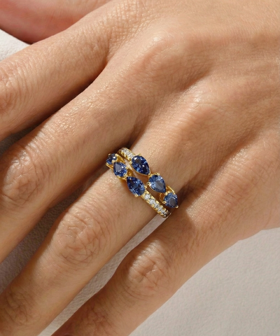 Gold ring with blue gemstones on a hand against a neutral background - alternate view