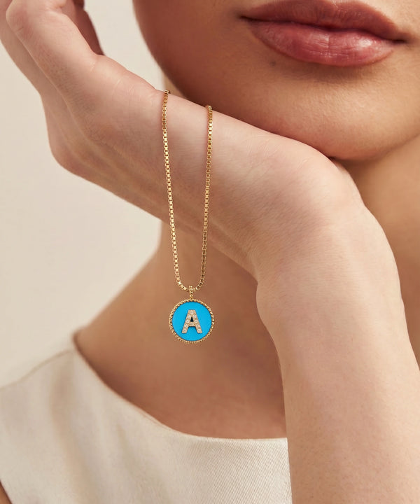 A close-up lifestyle shot of a model resting her chin on her hand, showcasing a delicate gold chain necklace with a circular pendant. The pendant features a bright blue enamel background with a white capital letter 'A' in the center, bordered by a gold bezel.