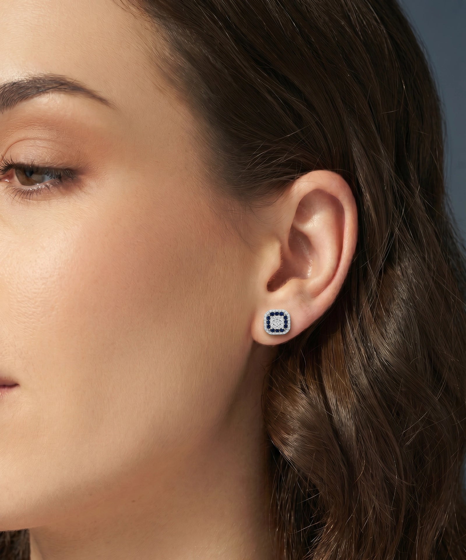 A close-up of a woman's ear wearing a small square stud earring with a silver-toned border and a central white stone surrounded by tiny blue stones.