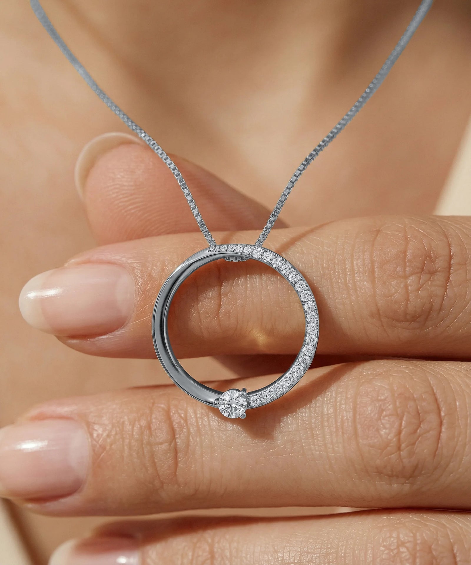 Silver necklace with a circular pendant held by a hand against a neutral background