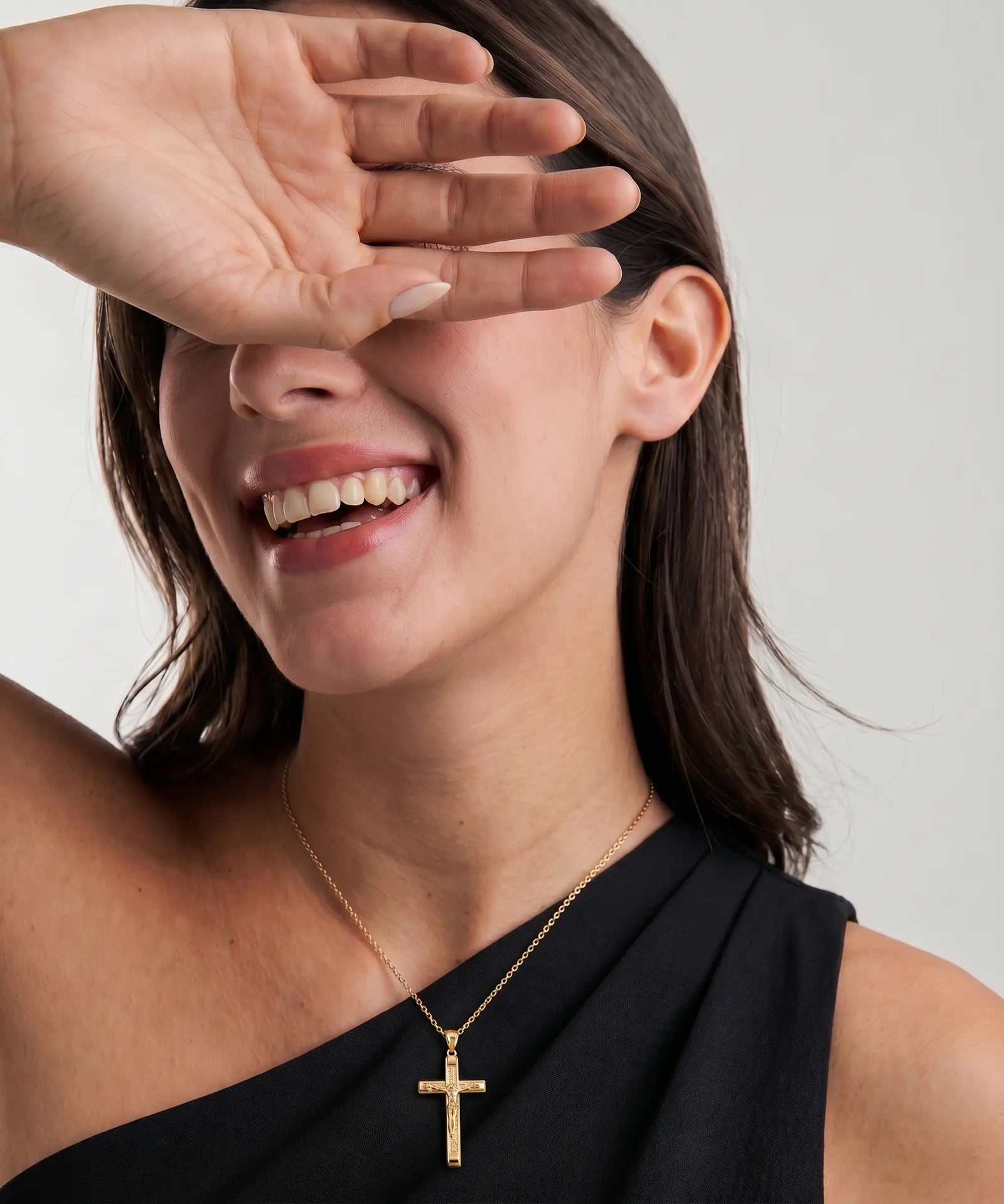 Woman wearing a gold necklace with a cross pendant, smiling and covering her eyes with one hand.