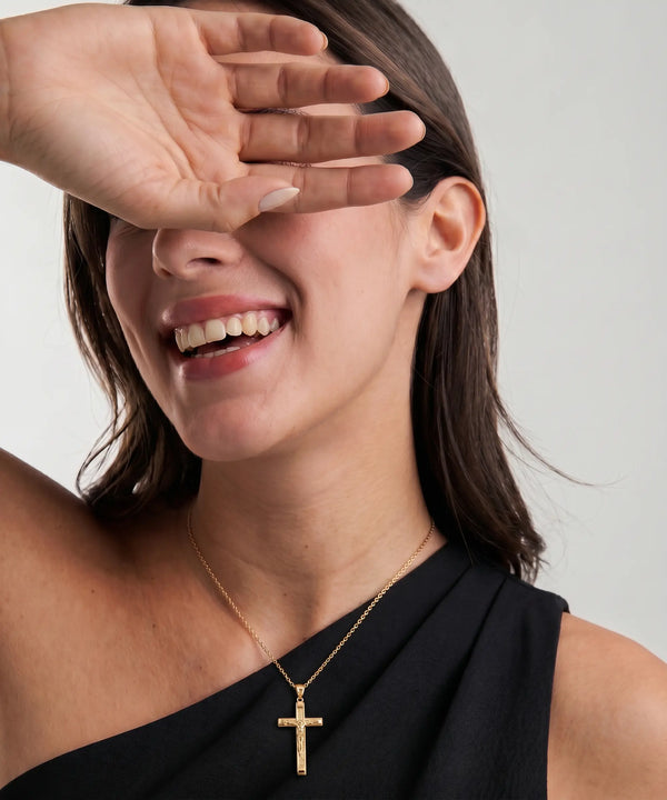 Woman wearing a gold necklace with a cross pendant, smiling and covering her eyes with one hand.