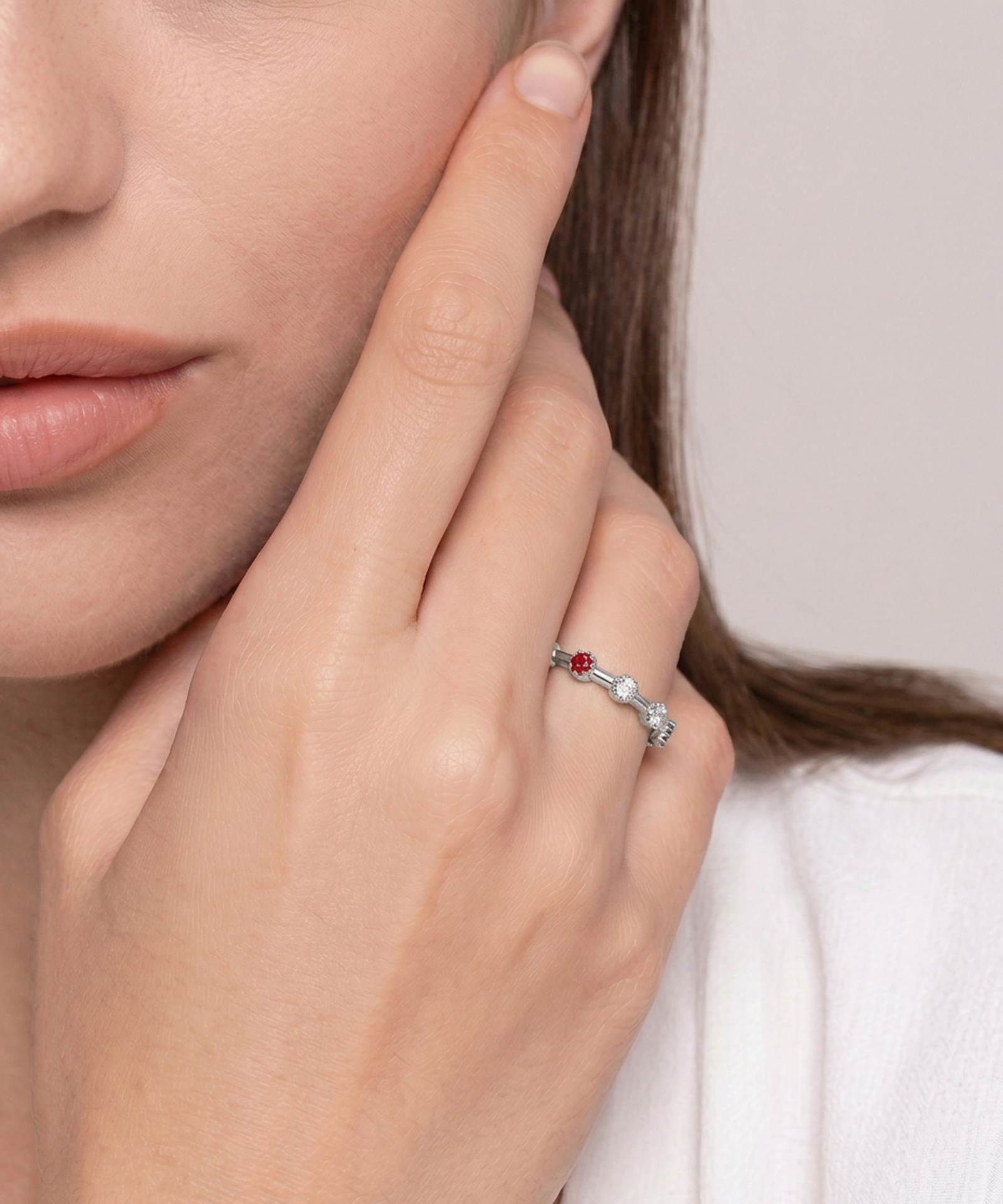 A close-up lifestyle shot of a woman's hand and face wearing a delicate silver ring with a small red gemstone and clear stones, set against a soft neutral background.