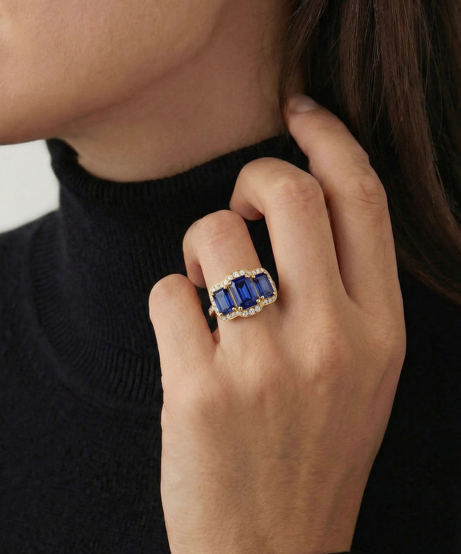 A close-up of a person wearing a statement ring with rectangular blue gemstones and small clear accent stones on a black turtleneck sweater.