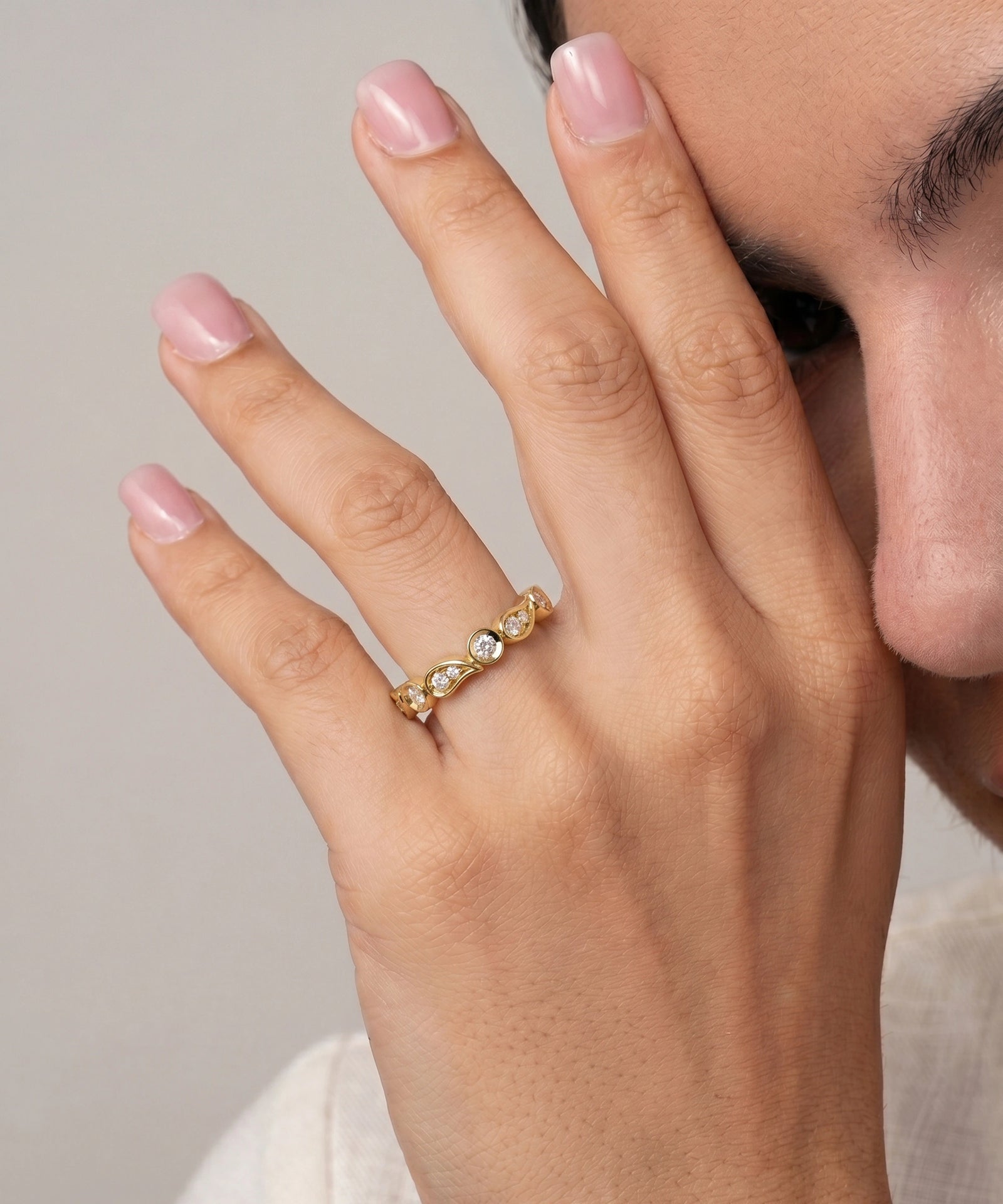 A close-up of a person's hand wearing a delicate gold band ring with three small clear gemstones, set against a soft neutral background.