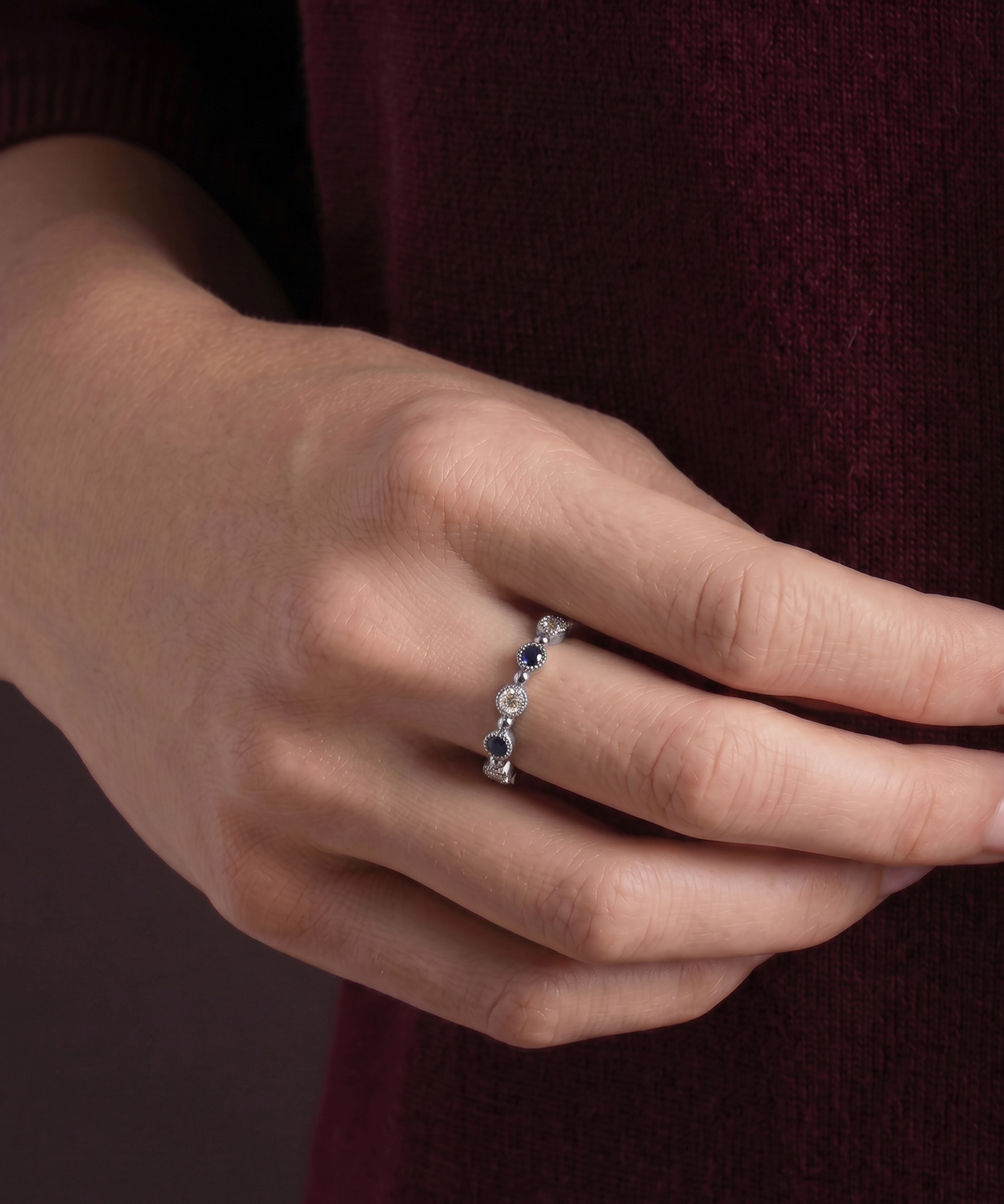 A close-up of a person's hand wearing a delicate silver ring with alternating dark blue and clear gemstones, set against a dark burgundy knit sweater.