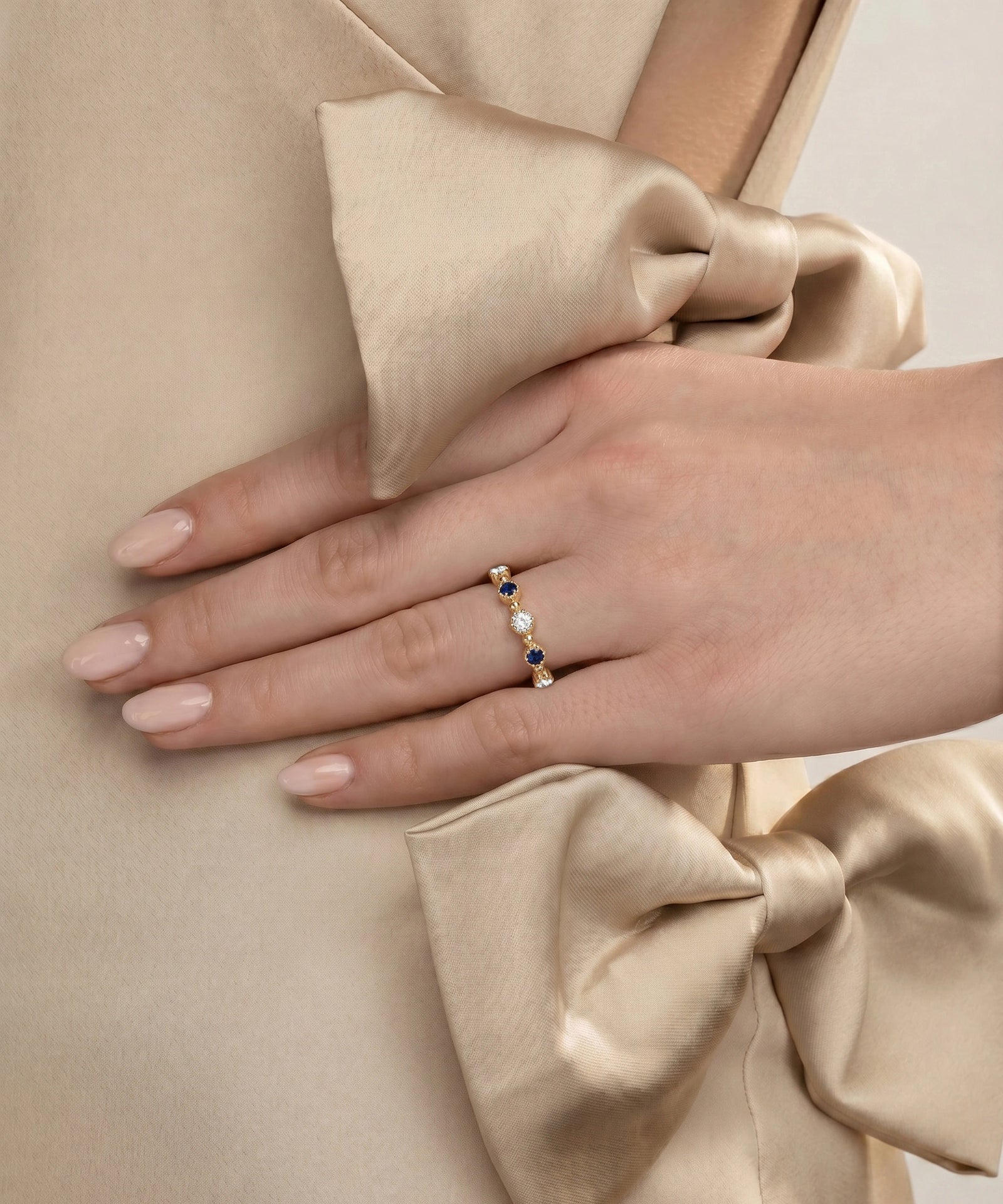 A close-up of a hand wearing a delicate gold ring adorned with small white pearls and dark blue gemstones, resting on a beige satin garment with a large bow.