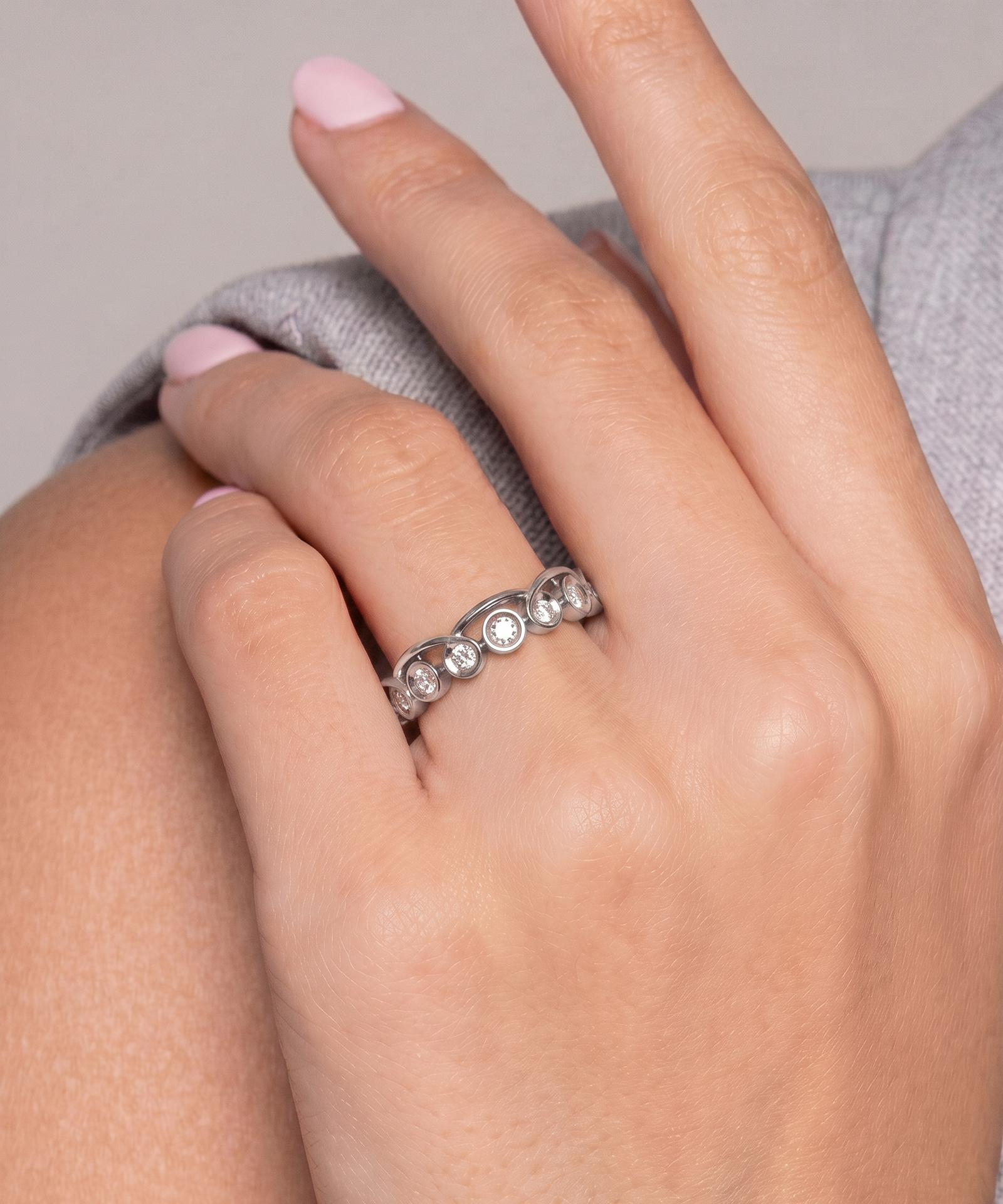 A close-up of a hand wearing a silver band ring with five clear round-cut gemstones, resting on a gray fabric surface.