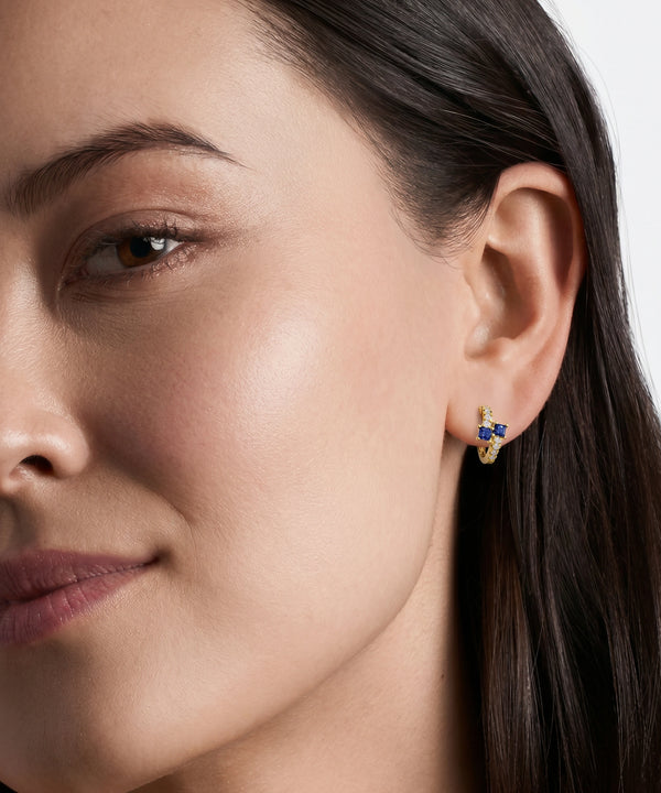 A close-up of a woman's ear wearing a small gold huggie hoop earring adorned with blue and white gemstones.