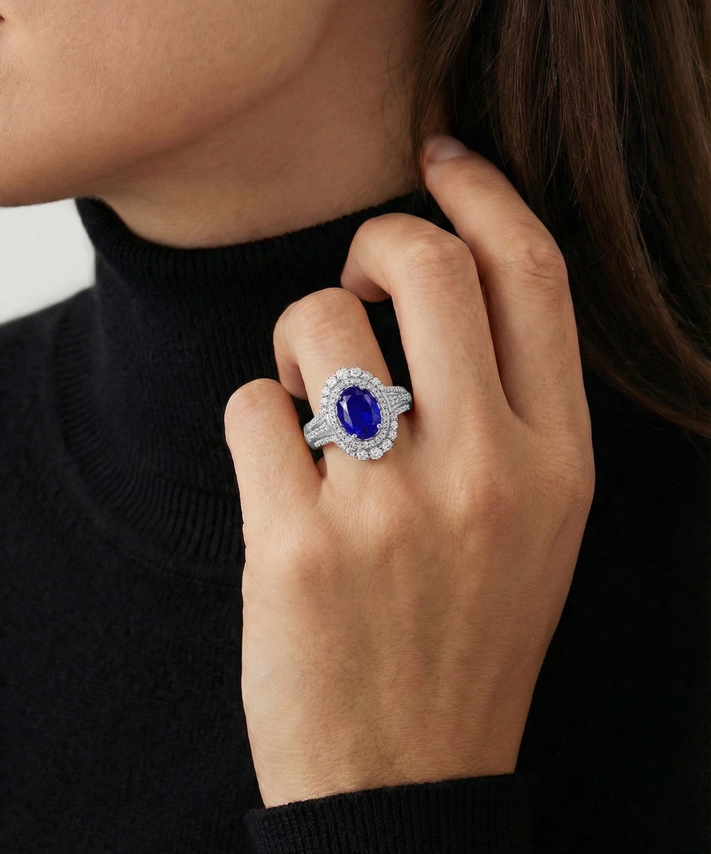 A close-up lifestyle shot of a model wearing a silver-toned halo ring with a large oval blue sapphire center stone and small accent diamonds.