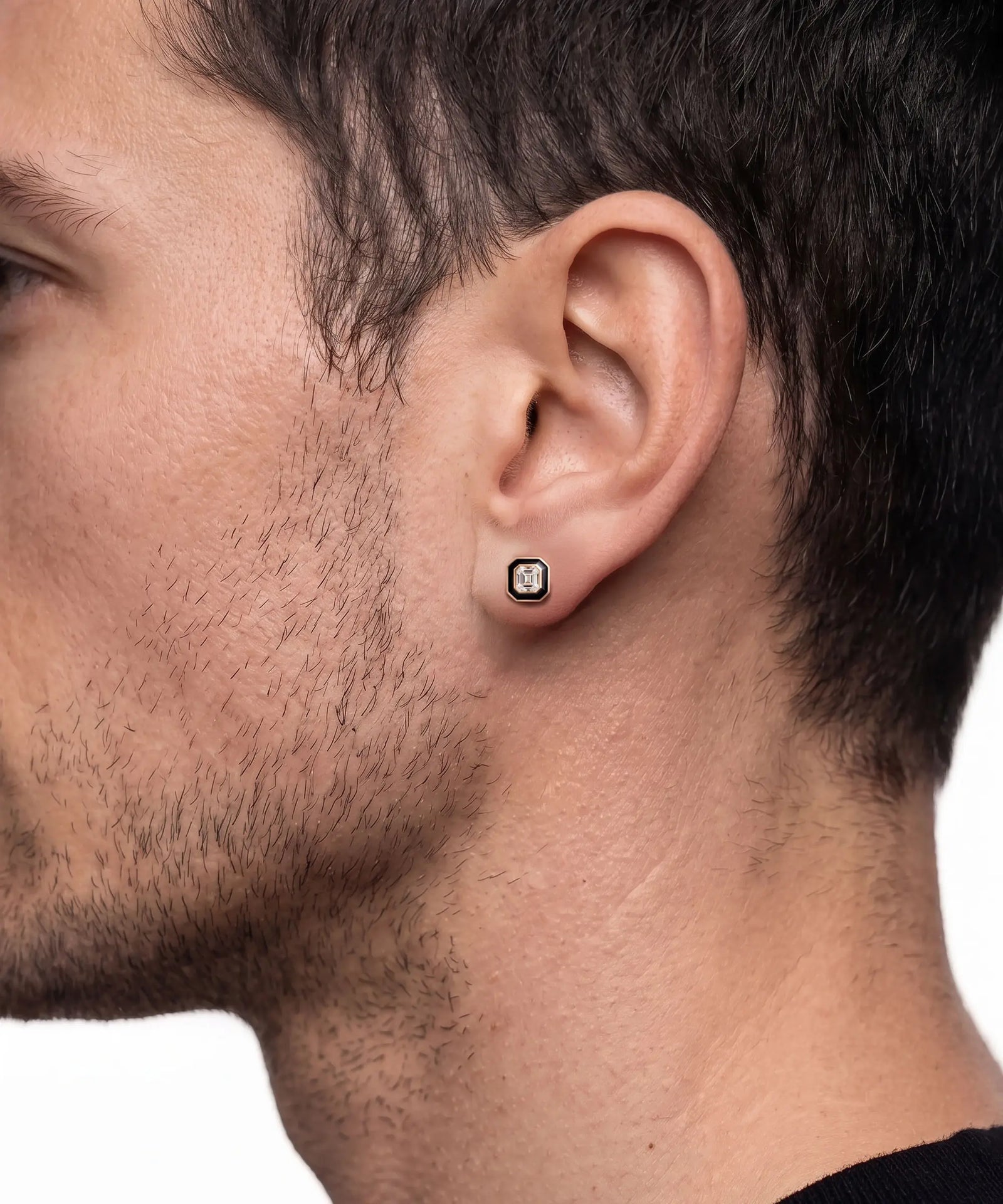 A close-up side profile of a man's head showing a small square stud earring with a black and white patterned center set in silver on his earlobe.