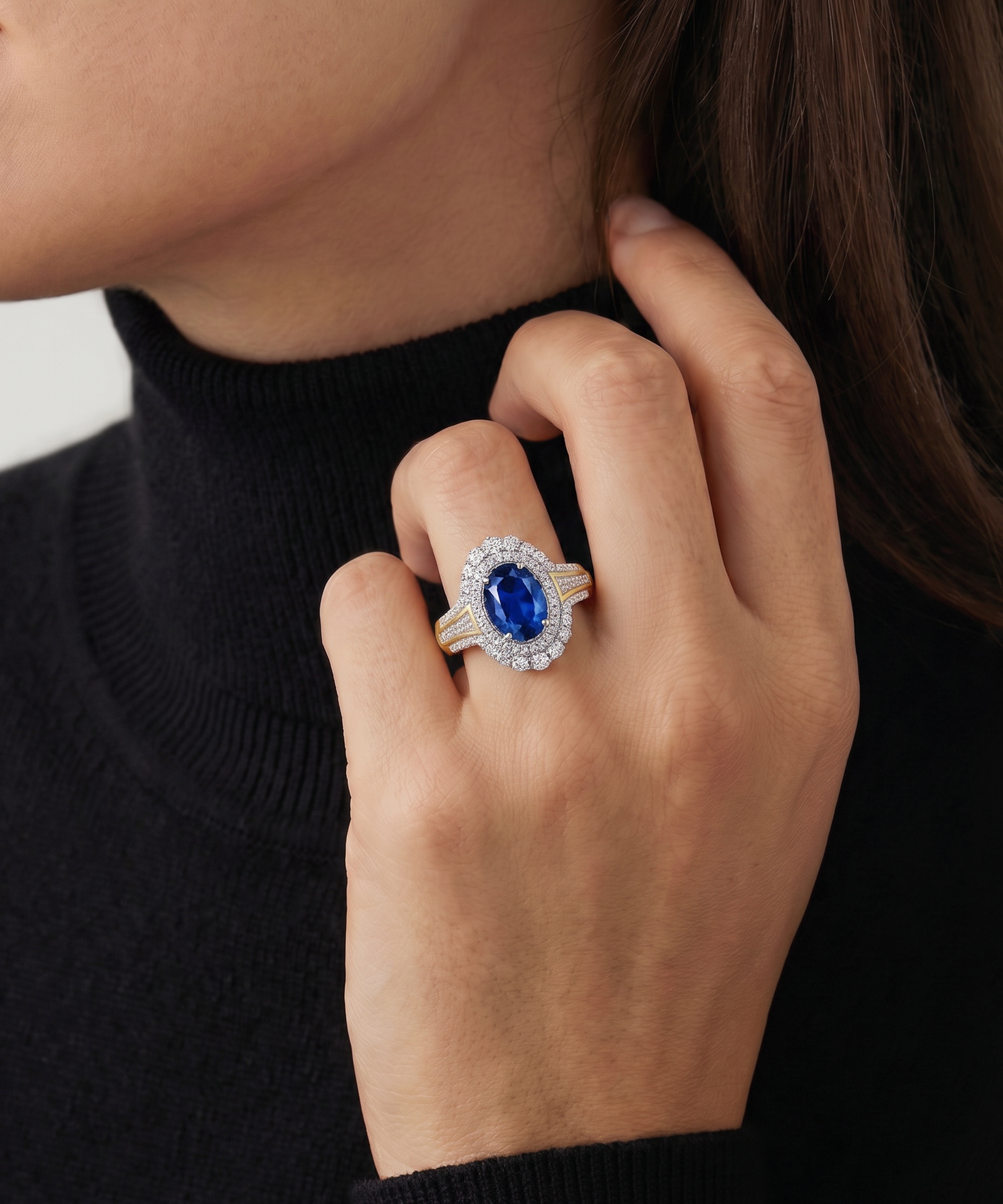 A close-up of a person's hand wearing a ring with a large oval blue gemstone surrounded by a halo of small clear stones, set on a black turtleneck sweater.
