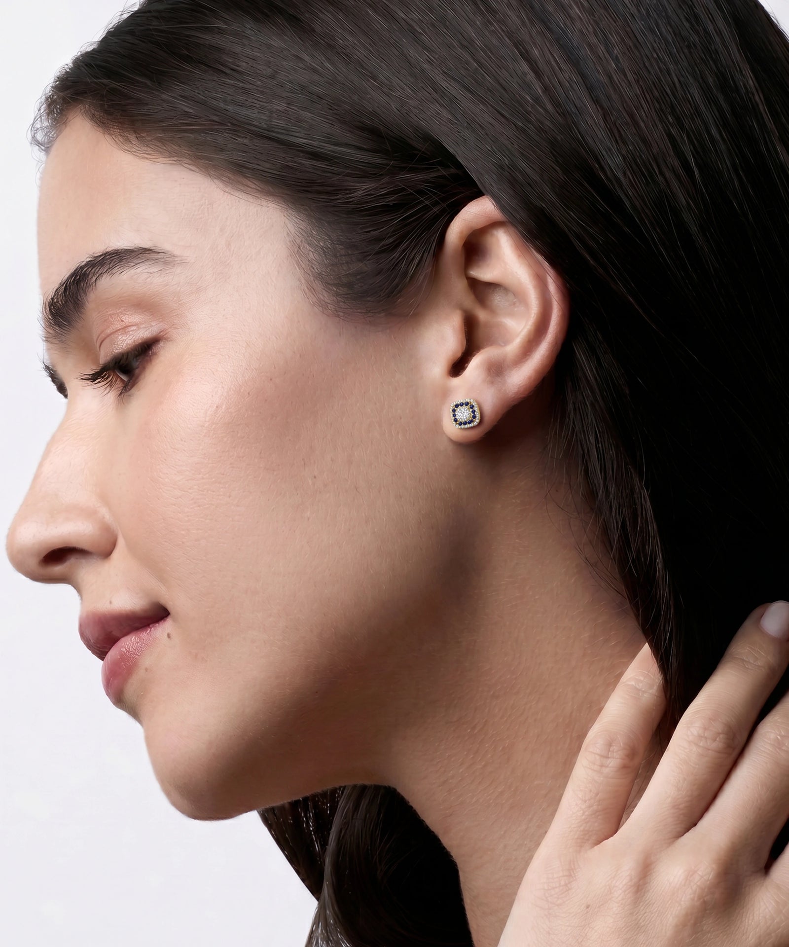 Close-up side profile of a woman wearing a small square stud earring with a silver-toned setting and clear stones, against a light gray background.