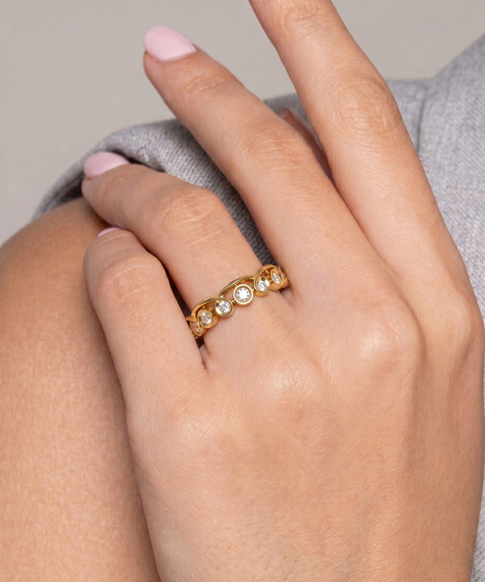 A close-up of a hand wearing a delicate gold band ring with small clear stones, resting against gray fabric.