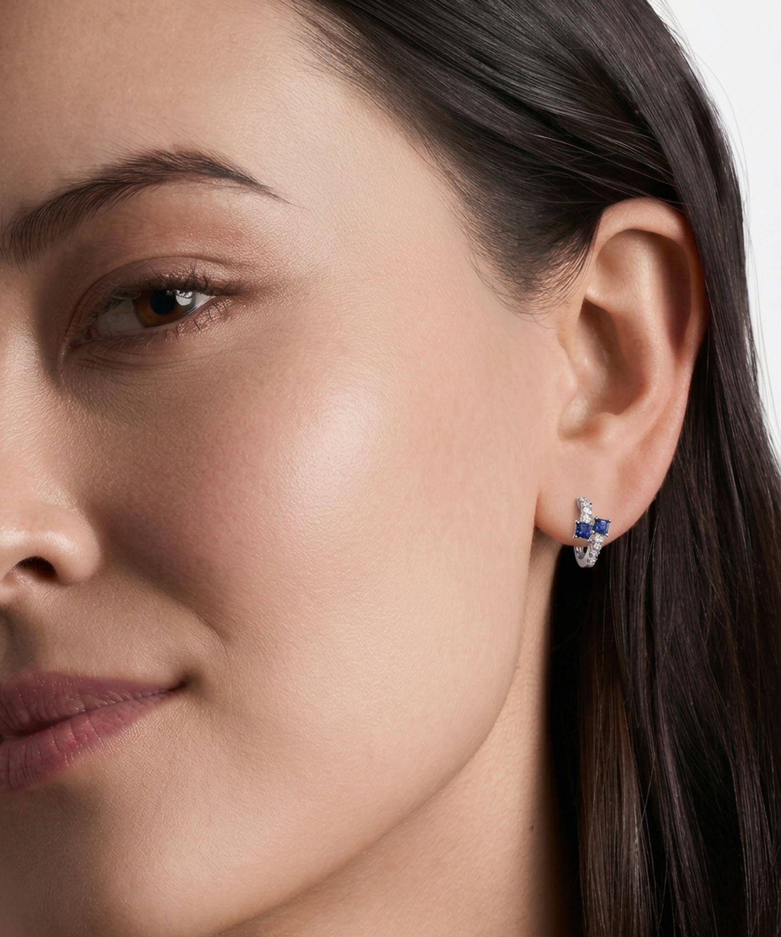 A close-up of a woman's ear wearing a small silver-toned earring with blue and clear gemstones, shown against a clean light background.